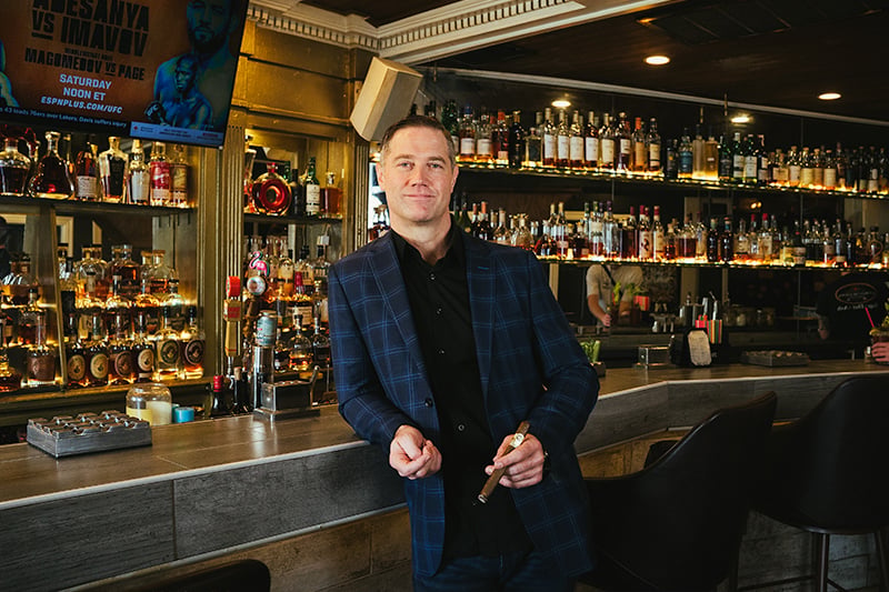 Cuban Creations owner Andrew Wilson holding a cigar at the bar, backed by the lounge's whiskey selection in New Orleans