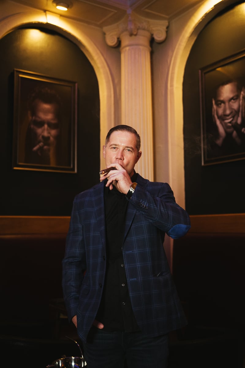 Cuban Creations owner Andrew Wilson smoking a cigar in the lounge beneath framed portraits and marble columns